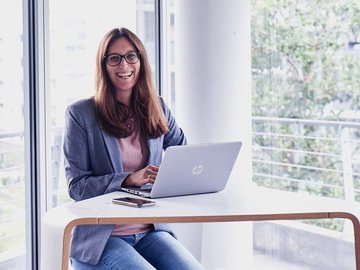 Smiling woman with long brown hair in an office sits in front of her laptop.